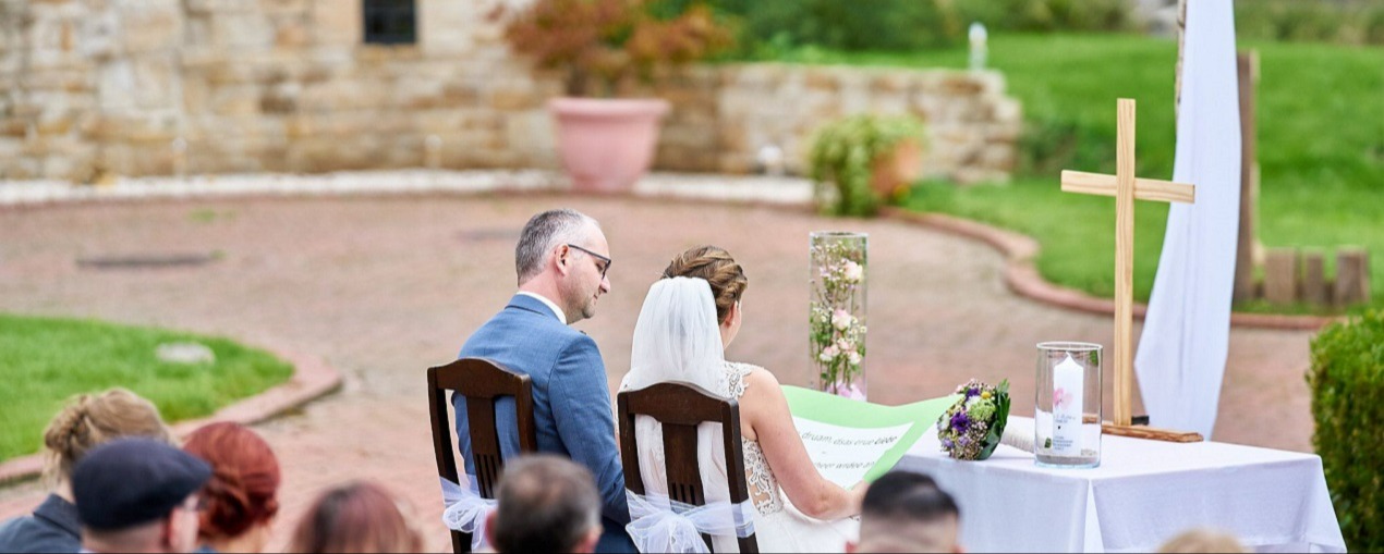 "Rückansicht eines Brautpaares bei der Hochzeitszeremonie im Freien: Die Braut im weißen Hochzeitskleid mit Schleier und der Bräutigam im blauen Anzug sitzen vor dem Traualtar mit Holzkreuz und Hochzeitskerze, während einer freien Trauung im Außenbereich des Kuckucks-Nests."