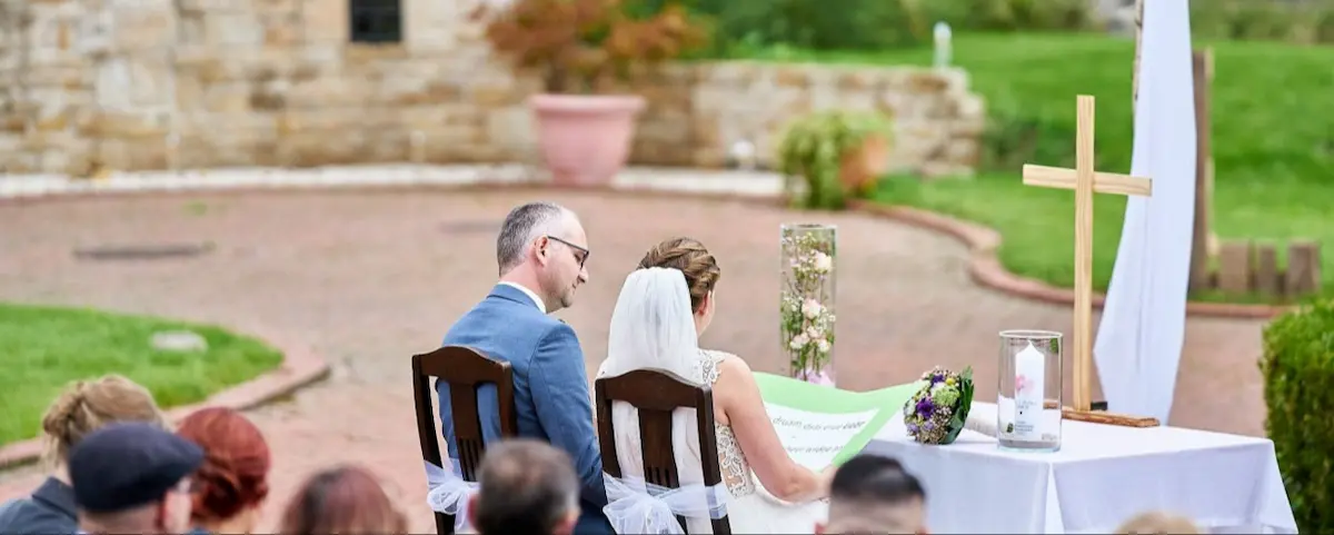 "Rückansicht eines Brautpaares bei der Hochzeitszeremonie im Freien: Die Braut im weißen Hochzeitskleid mit Schleier und der Bräutigam im blauen Anzug sitzen vor dem Traualtar mit Holzkreuz und Hochzeitskerze, während einer freien Trauung im Außenbereich des Kuckucks-Nests."