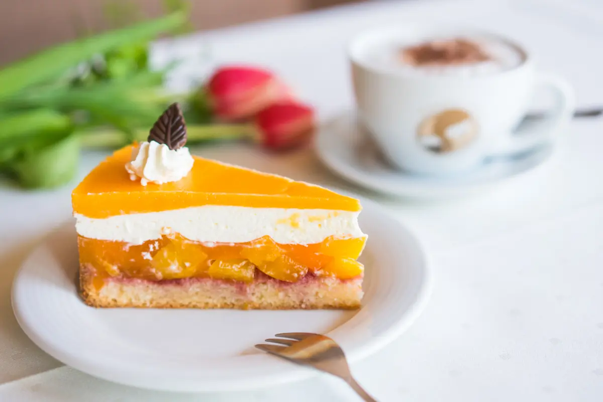 "Ein appetitliches Stück Pfirsich-Mandarinentorte mit Sahnehaube und Schokoblatt auf einem weißen Kaffeeteller. Im weichgezeichneten Hintergrund stehen eine Tasse Cappuccino mit Kakaopulver und frische rote Tulpen."