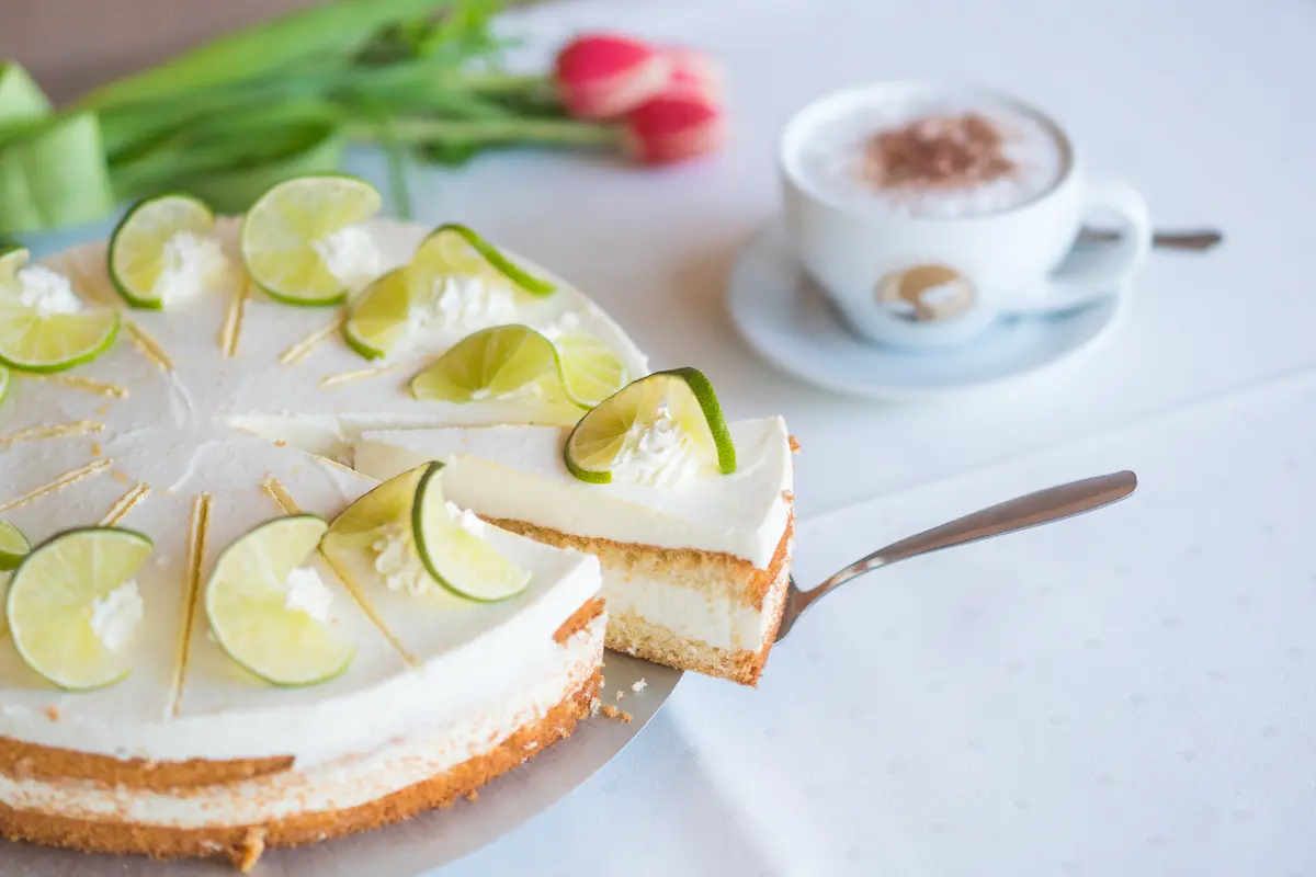 "Eine angeschnittene, cremige Limettentorte dekoriert mit Sahnetupfen und frischen Limettenscheiben. Ein Tortenheber hebt gerade ein Stück heraus. Im Hintergrund stehen eine Tasse Cappuccino und rote Tulpen."