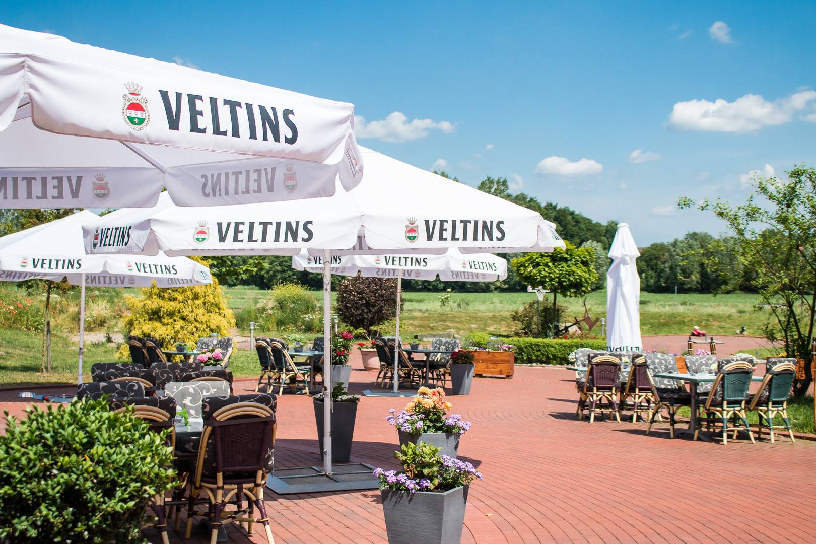 "Der einladende Biergarten des Eventhauses: Eine weitläufige Pflasterterrasse mit gemütlichen Tischen und Flechtstühlen unter großen, weißen Veltins-Sonnenschirmen. Im Hintergrund bietet sich ein freier Blick auf Wiesen und Bäume unter blauem Sommerhimmel."