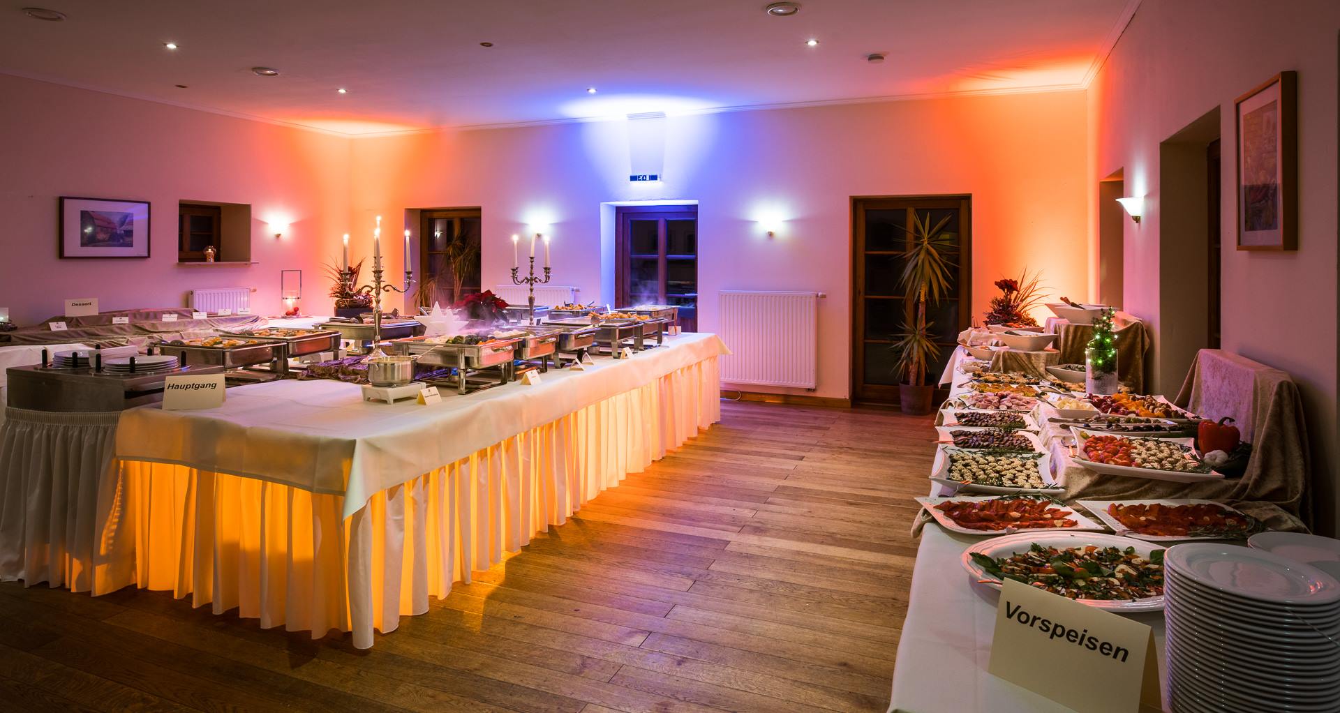 "Ein festlich aufgebautes Buffet in der Eventlocation: Auf der rechten Seite sind kalte Vorspeisen und Platten angerichtet, während im Hintergrund warme Hauptspeisen in Chafing-Dishes unter stimmungsvoller, farbiger Beleuchtung bereitstehen."