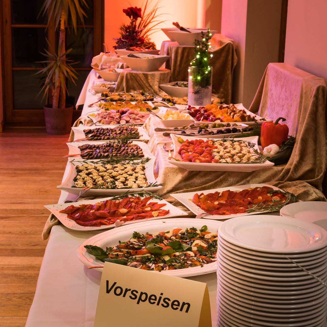 "Nahaufnahme des Buffets im Kuckucks-Nest: Eine lange Tafel mit verschiedenen kalten Vorspeisen wie gefüllten Tomaten, Antipasti-Platten, Salaten und Rouladen. Im Vordergrund steht ein Schild mit der Aufschrift 'Vorspeisen' neben einem Stapel weißer Teller."