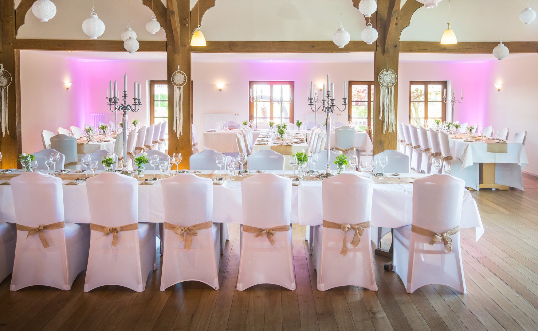 "Der festlich eingedeckte Festsaal im Kuckucks-Nest: Zu sehen sind mehrere lange Hochzeitstafeln mit weißen Stuhlhussen und Jutebändern auf rustikalem Parkettboden. Die Wände werden stimmungsvoll in Rosa beleuchtet, während von den hölzernen Deckenbalken weiße Papierlampions hängen."