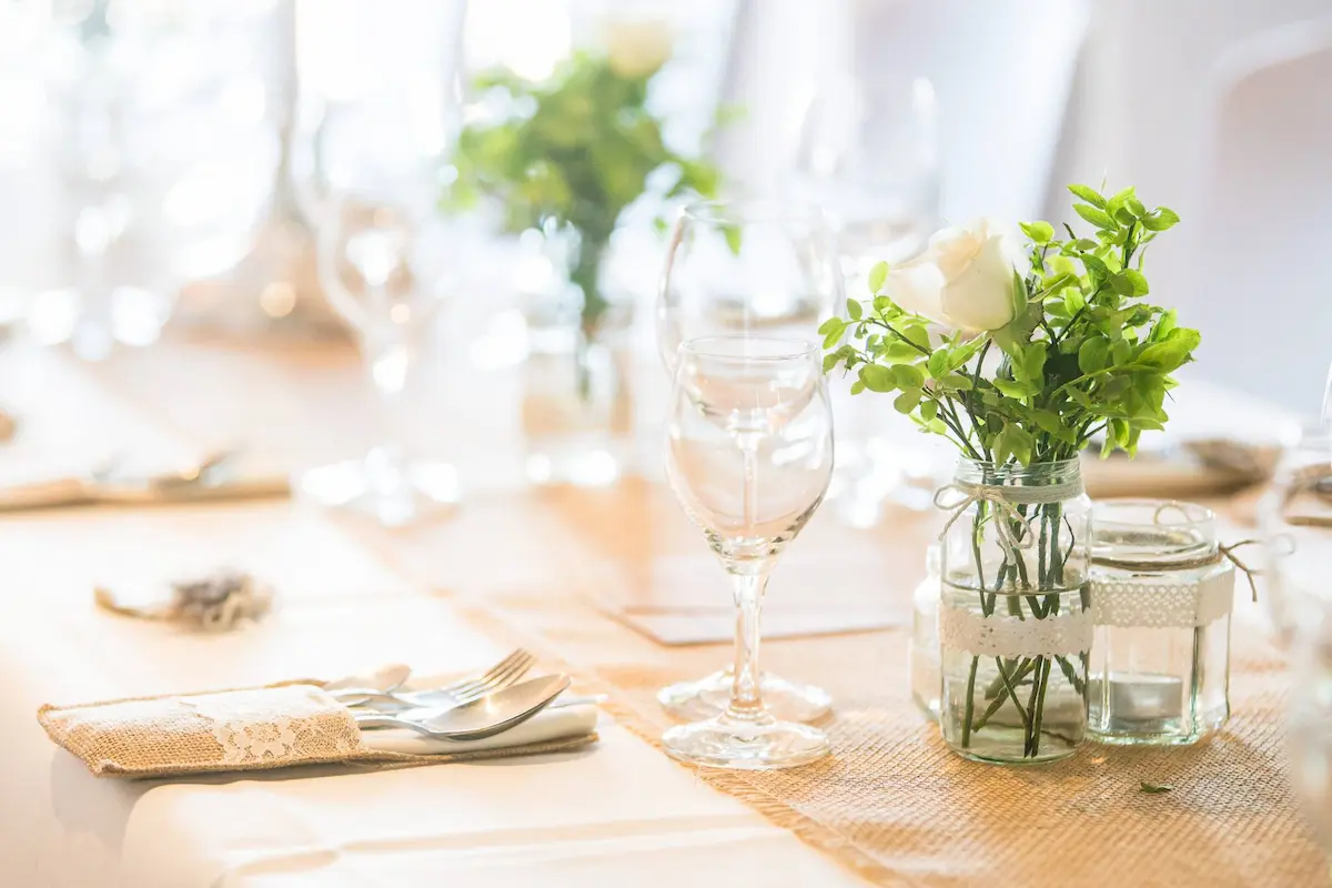 "Festliche Festtafel mit weißer Tischwäsche und einem Tischband aus Jute. Darauf stehen Weingläser, Bestecktaschen mit Spitzenborte und ein kleines Einmachglas mit einer weißen Rose und frischem Grün als Blumenschmuck."