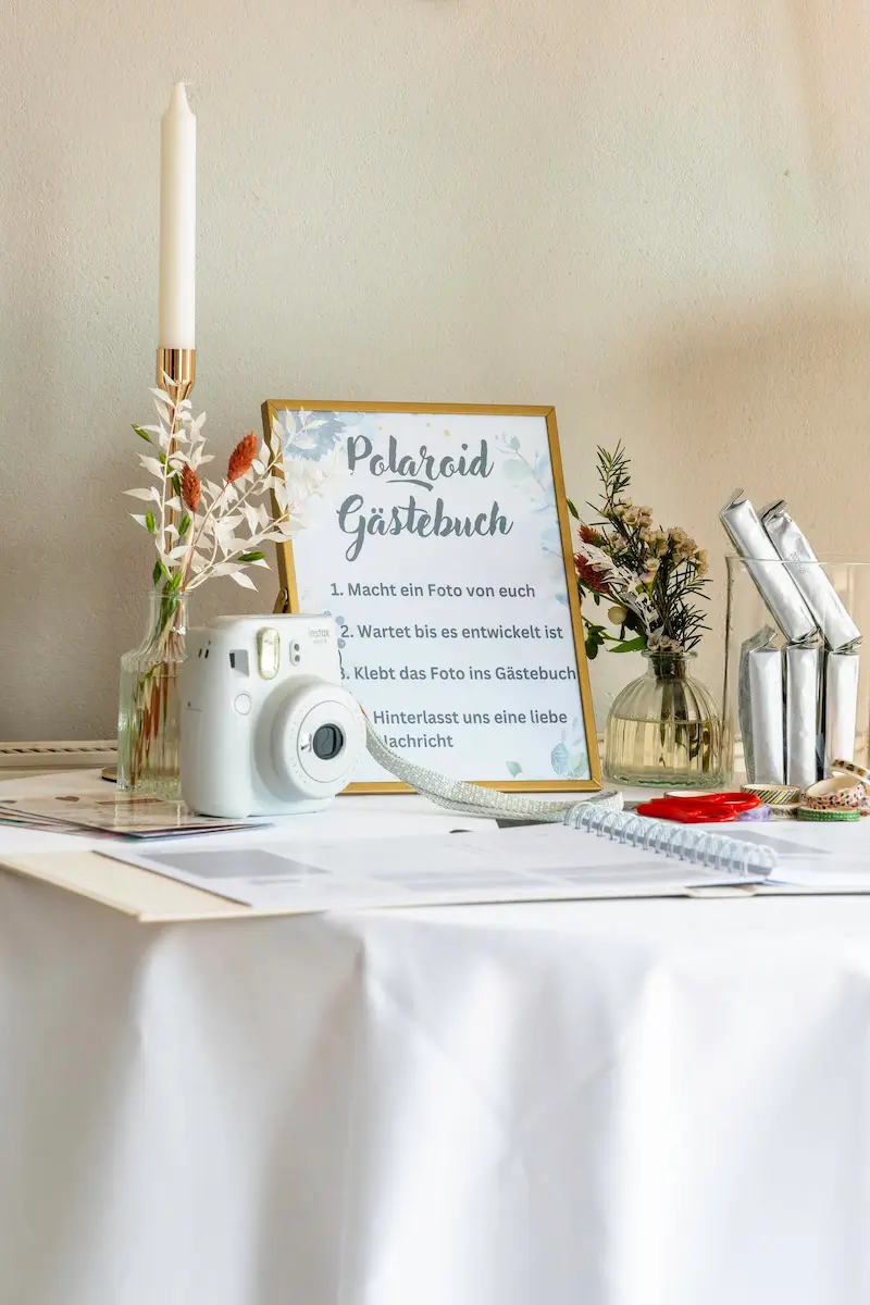 "Ein Hochzeitstisch mit einer weißen Instax-Sofortbildkamera und einem eingerahmten Schild mit der Aufschrift 'Polaroid Gästebuch'. Daneben stehen eine Stabkerze, kleine Blumenvasen und Bastelutensilien."