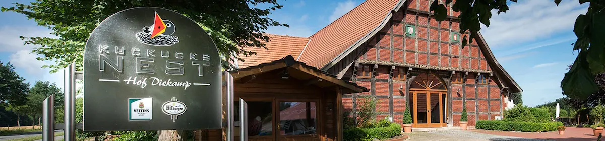 "Vorderansicht der Eventlocation: Im Vordergrund steht das dunkle Metall-Eingangsschild mit der Aufschrift 'Kuckucks Nest - Hof Diekamp'. Dahinter ist das große, restaurierte Fachwerkhaus mit rotem Ziegel-Mauerwerk und dem hölzernen Portal zu sehen."