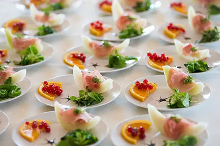 "Viele liebevoll angerichtete Vorspeisenteller auf einem Buffet im Kuckucks-Nest: Jedes Tellerchen ist garniert mit einer Melonenspalte und Schinken, etwas Feldsalat sowie einer Orangenscheibe mit frischen roten Johannisbeeren."