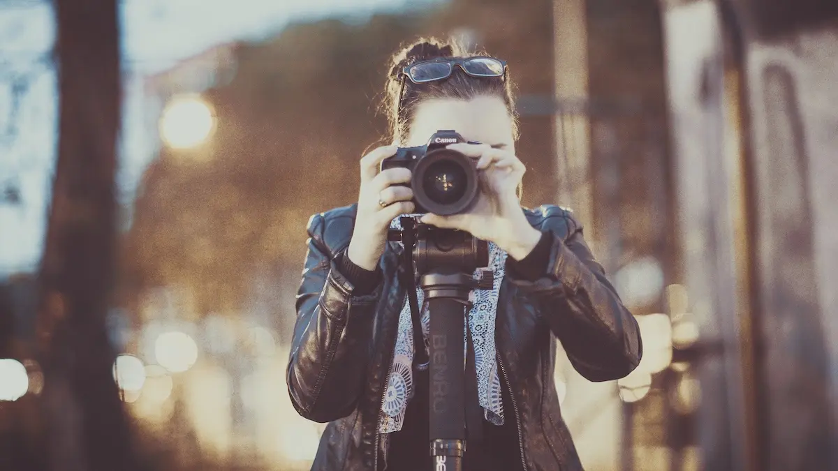 "Nahaufnahme einer jungen Fotografin im Kuckucks-Nest, die mit einer Canon-Spiegelreflexkamera auf einem Stativ ein Foto macht. Im Hintergrund schimmert weiches Licht (Bokeh-Effekt)."