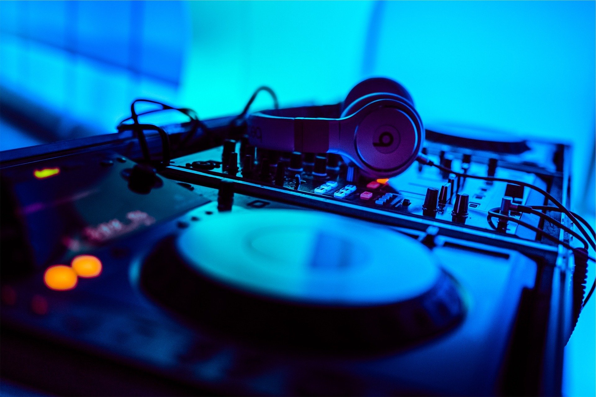 "Nahaufnahme eines DJ-Decks im Kuckucks-Nest: Ein Mischpult mit darauf abgelegten Kopfhörern, beleuchtet von Club-Atmosphäre in kräftigem Blau mit orange leuchtenden Knöpfen im Vordergrund."